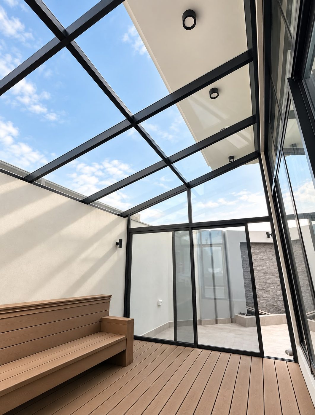 Black aluminium glass roof and sliding glass doors enclosing a bright courtyard with a wooden deck