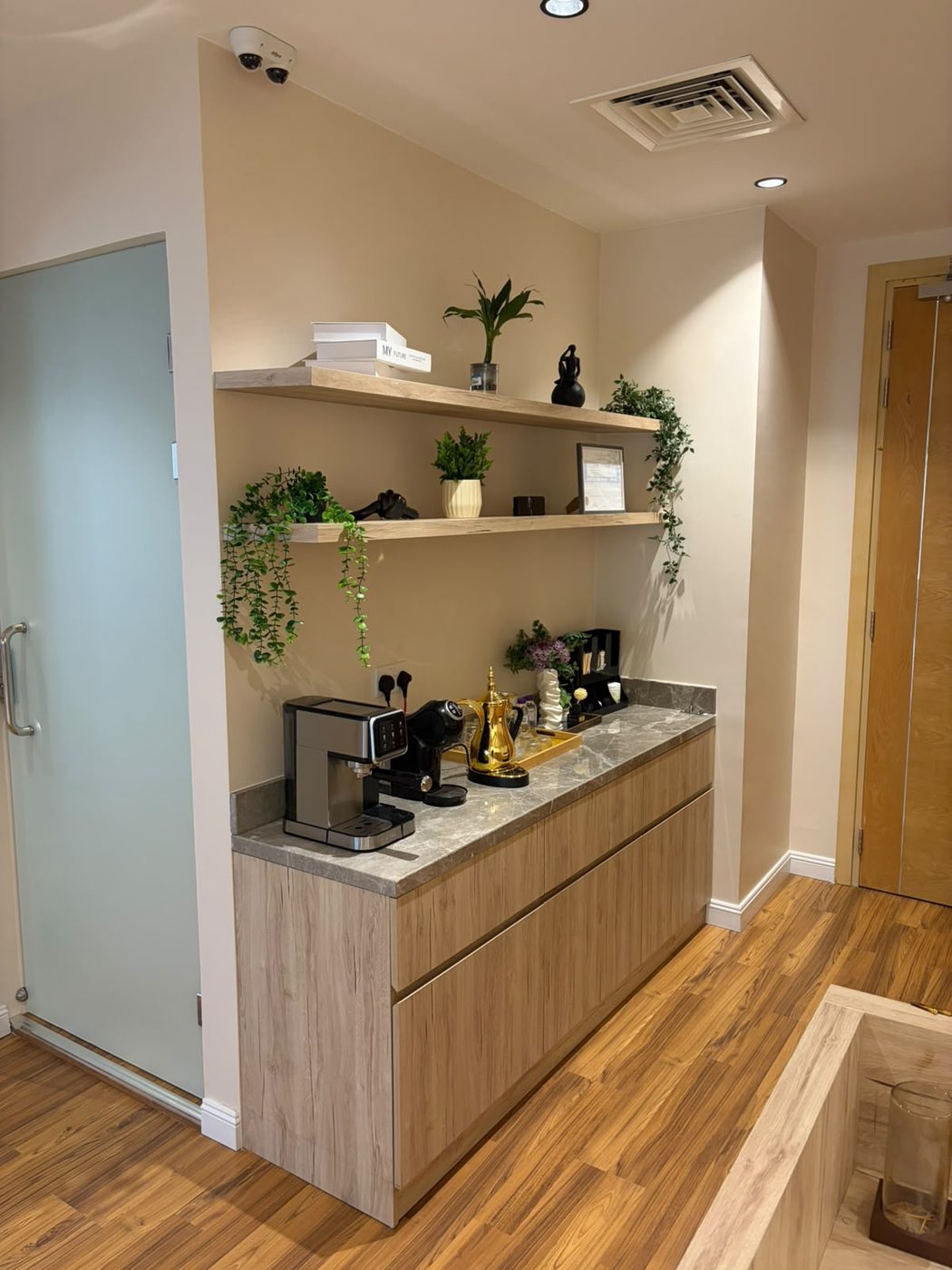 Office pantry corner with light-oak cabinets and a grey stone worktop set between a frosted glass door and a timber door