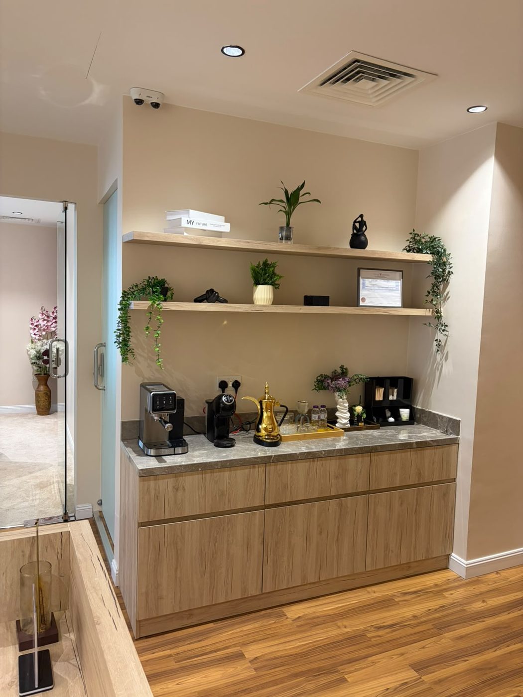 Side view of an office coffee station with light-oak cabinets, a grey stone worktop and floating shelves beside a glass door